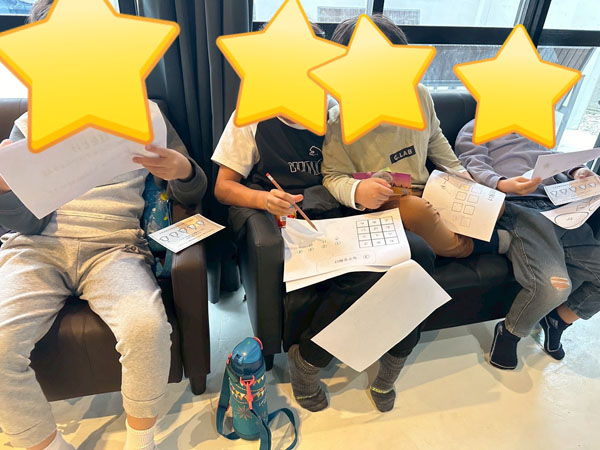 放課後等デイサービスの謎解きイベントの様子。児童達がソファに座り、紙に描かれた謎を解いています。